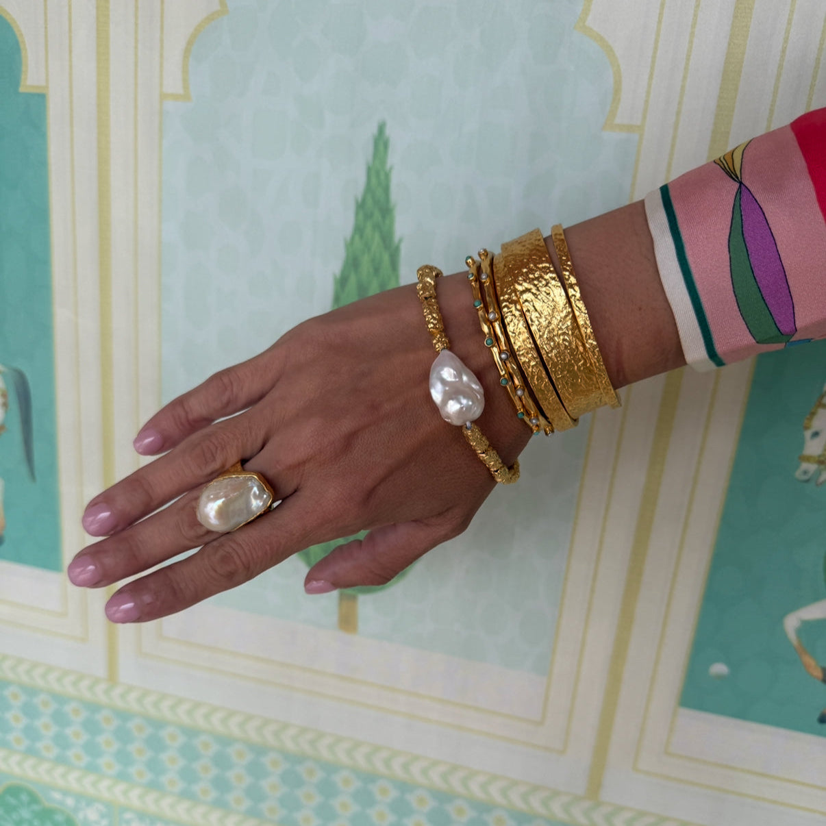 Hand wearing gold bangles and a ring on a patterned background